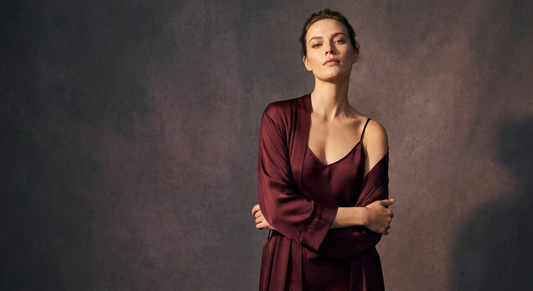 A confident woman stands in dramatic studio lighting wearing rich burgundy silk lingerie, her posture tall and empowered against a warm shadowed background.