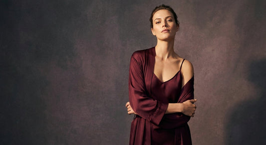 A confident woman stands in dramatic studio lighting wearing rich burgundy silk lingerie, her posture tall and empowered against a warm shadowed background.